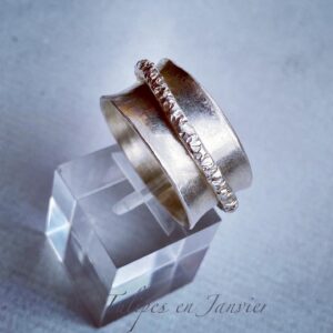 Anneau moyennement large en argent, entouré d'un anneau fin avec une forte texture martelée, posé sur un cube transparent sur un fond gris