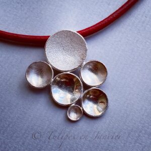 Un pendentif argenté composé de 6 disques concaves de tailles variées monté sur un cordon de cuir rouge.