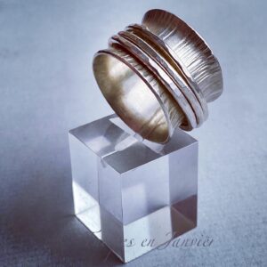 Bague large ornée d'un motif de stries et de trois anneaux finement martelés, posée sur un cube transparent sur un fond gris.