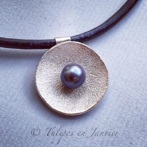 Un disque concave avec une légère texture est orné d'une perle violet foncé au milieu. Le tout est monté sur un cordon noir.