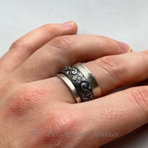 Une main qui porte un anneau large en argent sur le majeur. L'anneau est en argent et a un deuxième anneau légèrement plus fin avec des motifs floraux avec des volutes.