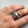 Une main porte un large anneau argenté à son majeur. L'anneau a des stries verticales et un deuxième anneau plus fin, décoré de motifs floraux, l'entoure.