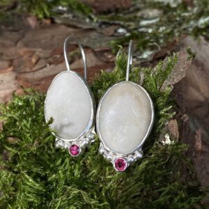 Une paire de boucles d'oreille faites de galets ovales gris clair, avec un sertissage argenté et une petite pierre rose sous les galets, encadrée par des petits granules en argent. Le tout est posé sur un morceau d'écorce d'arbre entouré de mousse.